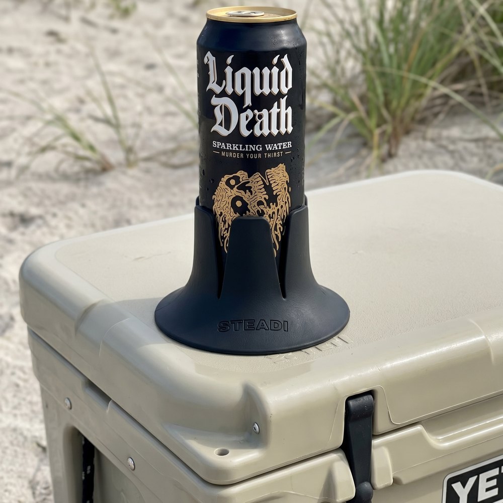 A tall can standing in a black Steadi on the lid of a hard cooler, with sand and dune grass behind.